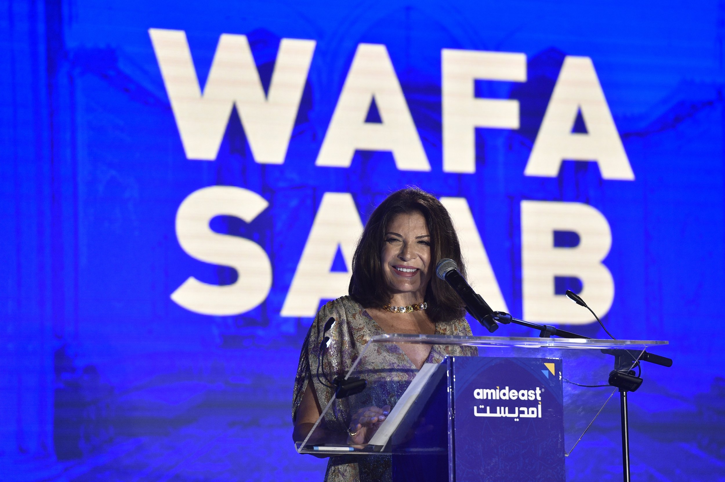 Wafa Saab smiles while speaking at a podium with the Amideast logo. Her name is displayed prominently on a screen behind her.
