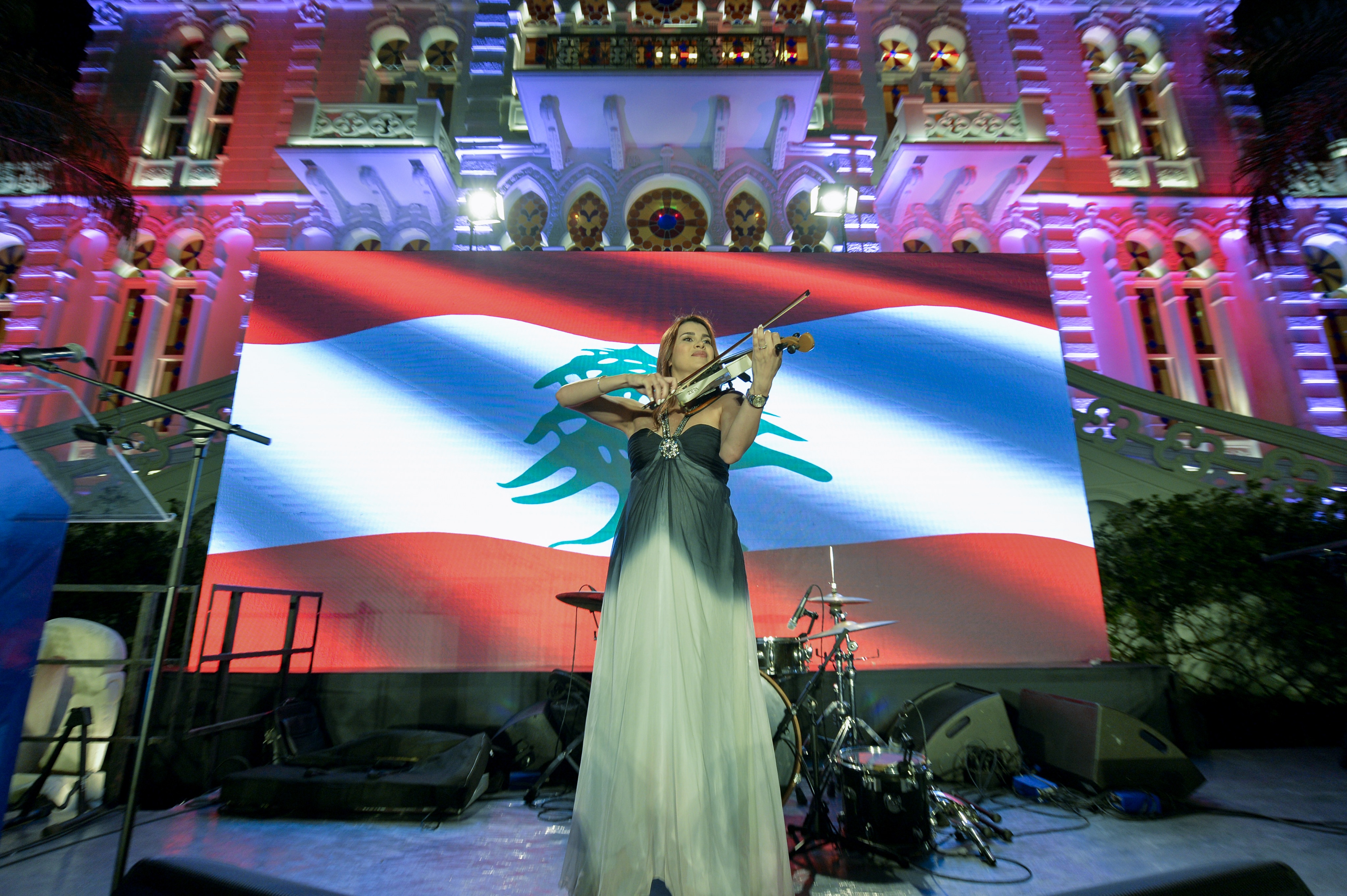 A woman in an elegant gown plays the violin on stage, with the Lebanese flag displayed on a screen behind her and an illuminated building as the backdrop.
